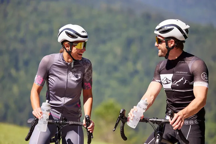 Zwei Rennradfahrer mit Helm machen lachend eine Pause vor grüner Berglandschaft