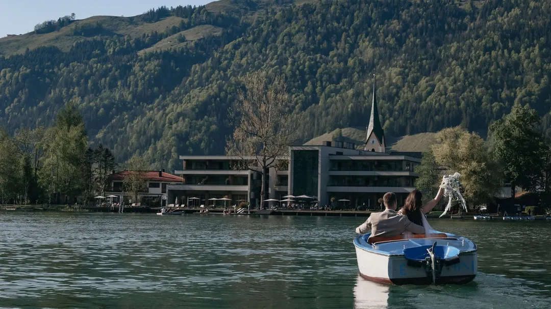 Brautpaar im Ruderboot auf einem See vor Hotel und Bergkulisse