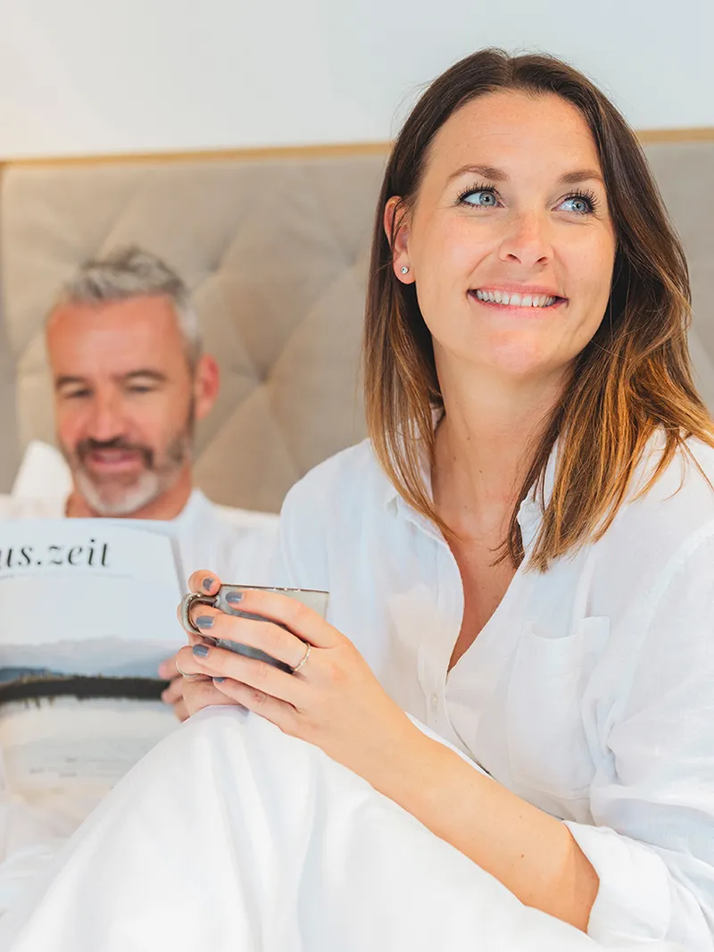 Glückliches Paar entspannt im Bett. Sie lächelt mit einer Tasse, er liest "aus.zeit". Bequeme, weiße Bettwäsche.