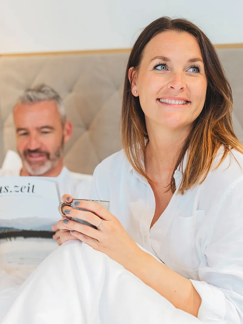 Glückliches Paar entspannt im Bett. Sie lächelt mit einer Tasse, er liest "aus.zeit". Bequeme, weiße Bettwäsche.
