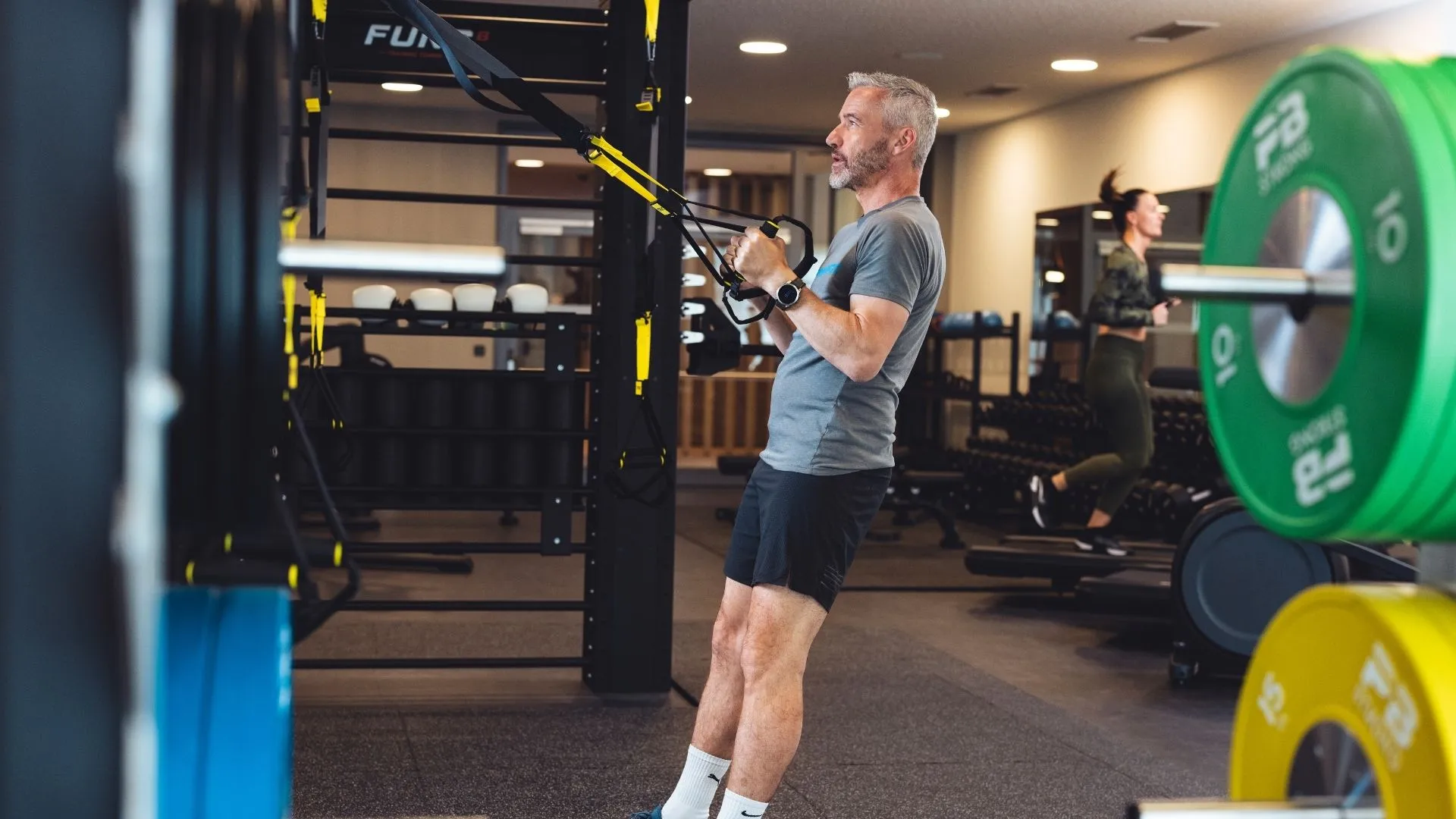 Mann trainiert mit Schlingentrainer im Fitnessstudio, im Hintergrund Frau auf Laufband