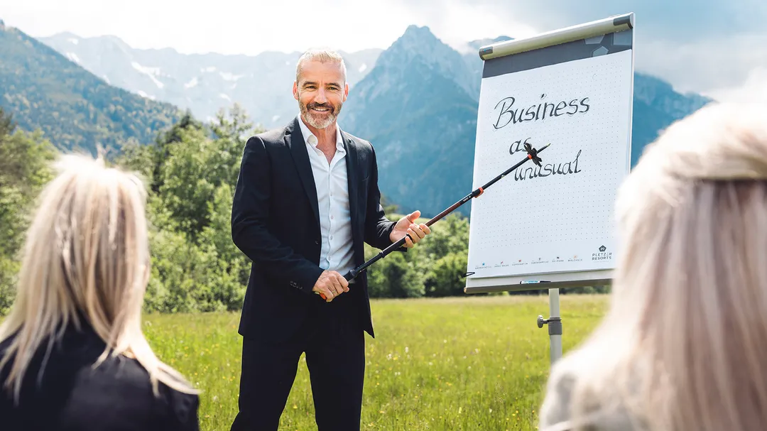 Ein lächelnder Mann im Anzug präsentiert im Freien vor einem Flipchart mit "Business as unusual" und Bergpanorama vor zwei Zuhörerinnen.