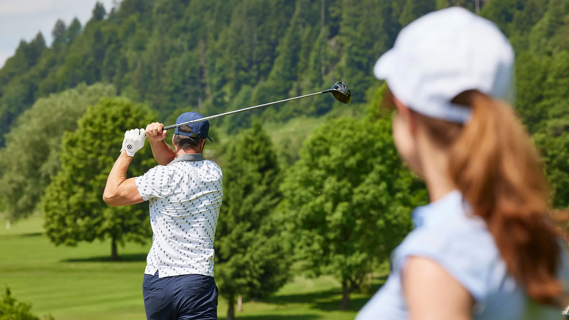 Golfer beim Abschlag auf einem Golfplatz, im Vordergrund eine zuschauende Frau