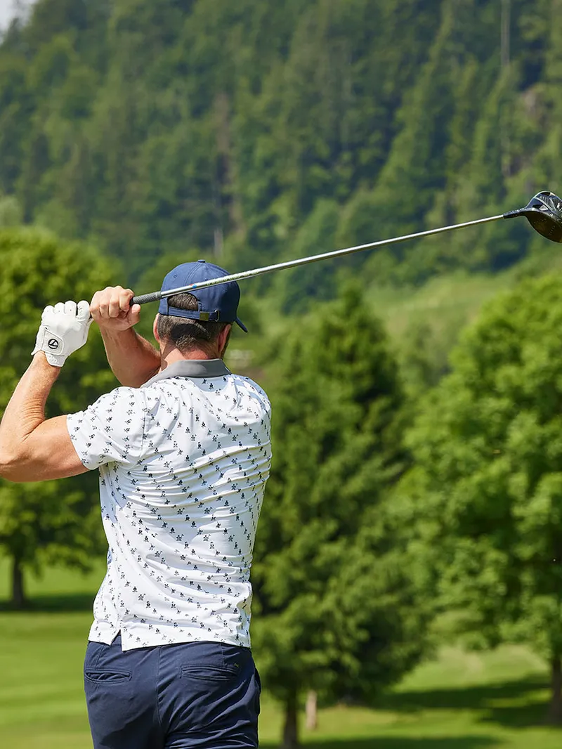 Golfer beim Abschlag auf einem Golfplatz, im Vordergrund eine zuschauende Frau