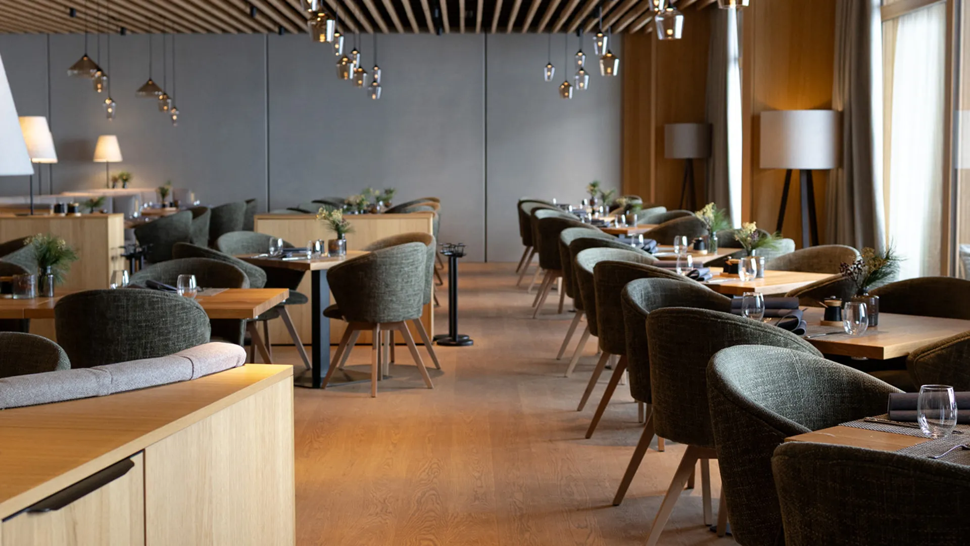 Modernes Restaurant mit Holzböden, gedeckten Tischen, dunkelgrünen Stühlen, einer Lamellenholzdecke und stilvollen Pendelleuchten.