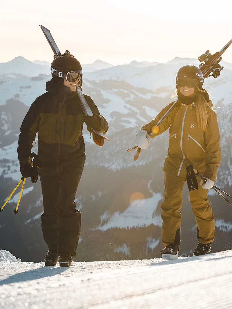 Zwei Skifahrer mit Skiern über der Schulter auf einer Piste vor Bergpanorama