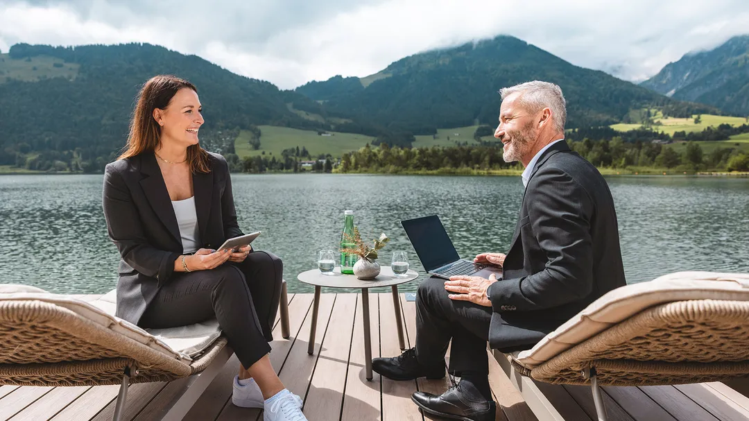 Zwei Geschäftsleute lächeln sich auf Liegestühlen an einem malerischen See mit Bergen im Hintergrund an, arbeiten mit Laptop und Tablet.