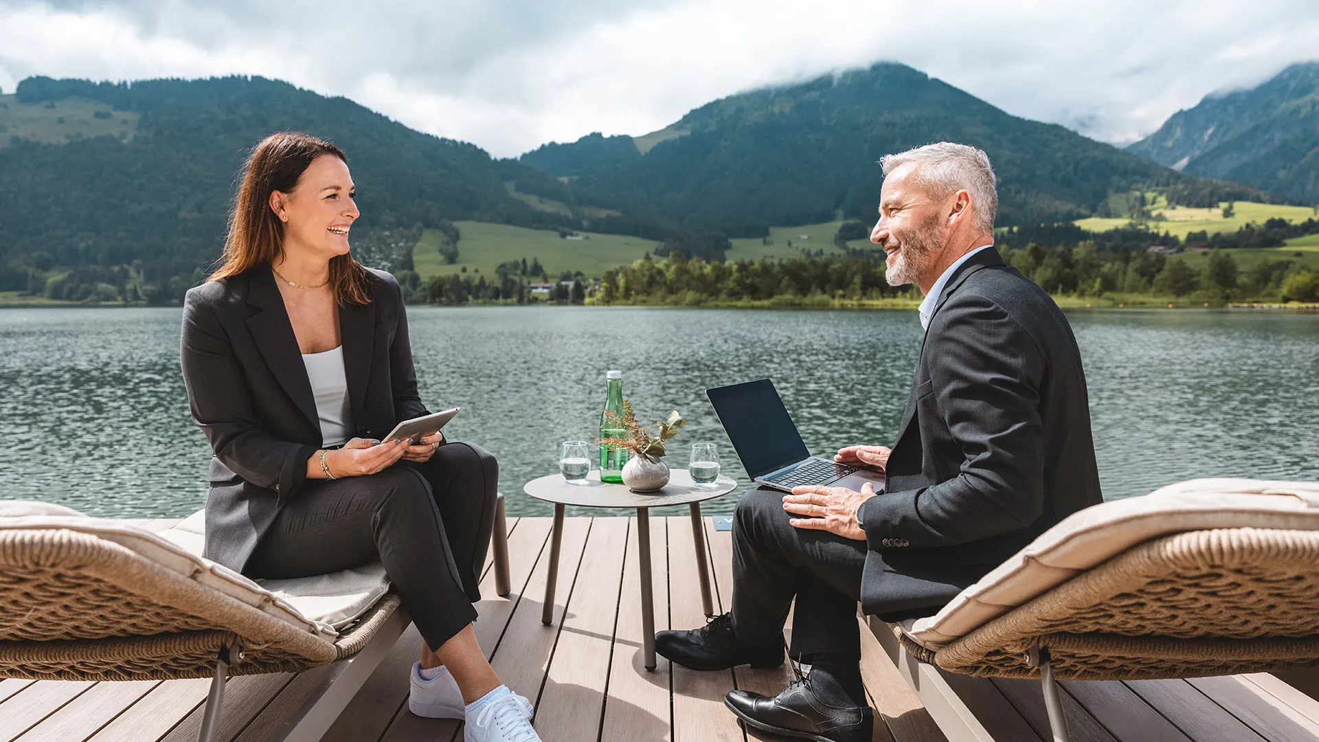 Zwei Geschäftsleute lächeln sich auf Liegestühlen an einem malerischen See mit Bergen im Hintergrund an, arbeiten mit Laptop und Tablet.