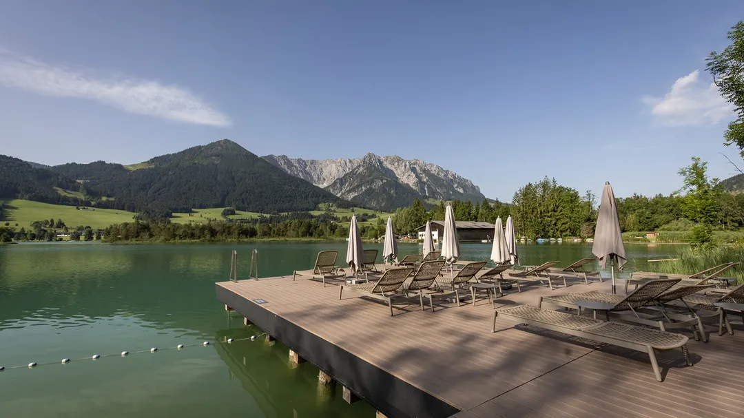 Ein Holzsteg mit Liegestühlen und Sonnenschirmen am grünen See vor bewaldeten Hügeln und majestätischen Bergen unter blauem Himmel.