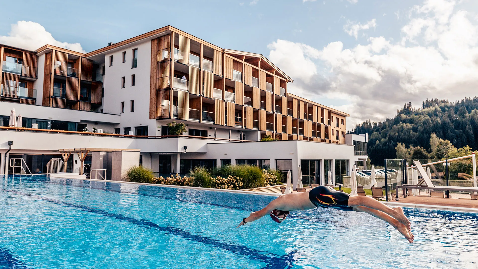 Mann springt in den Außenpool vor einem Hotelgebäude mit Holzfassade