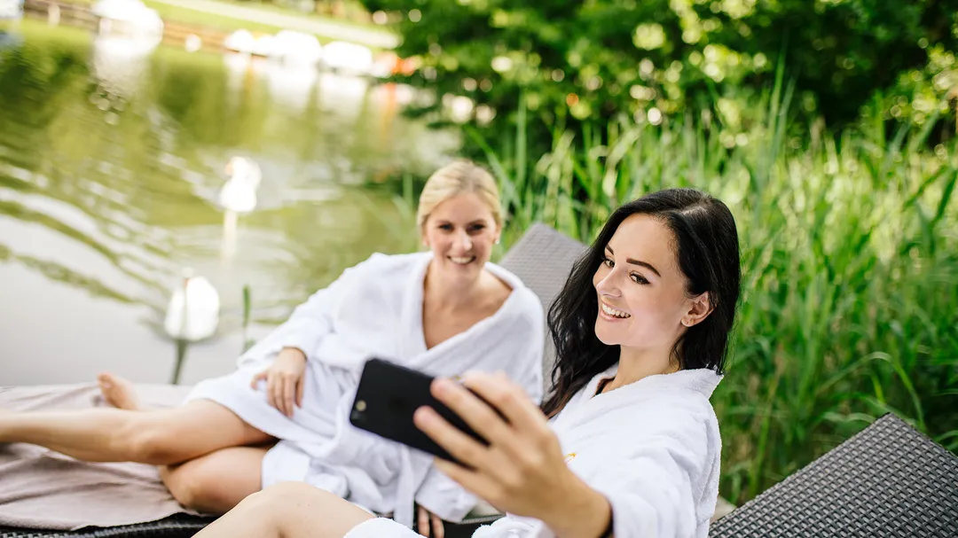 Zwei Frauen im Bademantel machen ein Selfie auf Liegestühlen am See