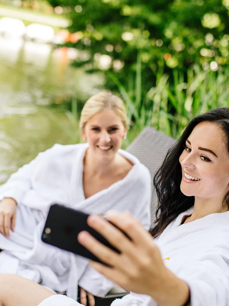 Zwei Frauen im Bademantel machen ein Selfie auf Liegestühlen am See