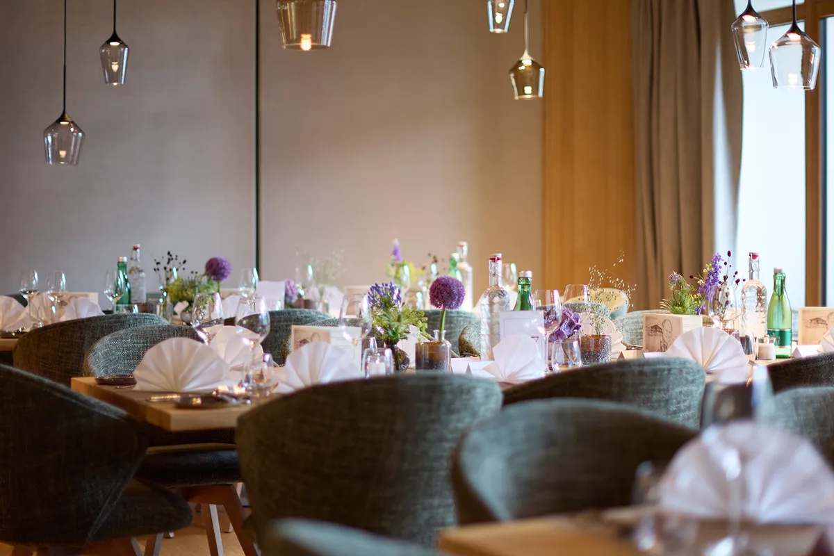 Restaurant mit festlich gedeckten Holztischen, grünen Stühlen, Blumenarrangements und modernen Pendelleuchten.