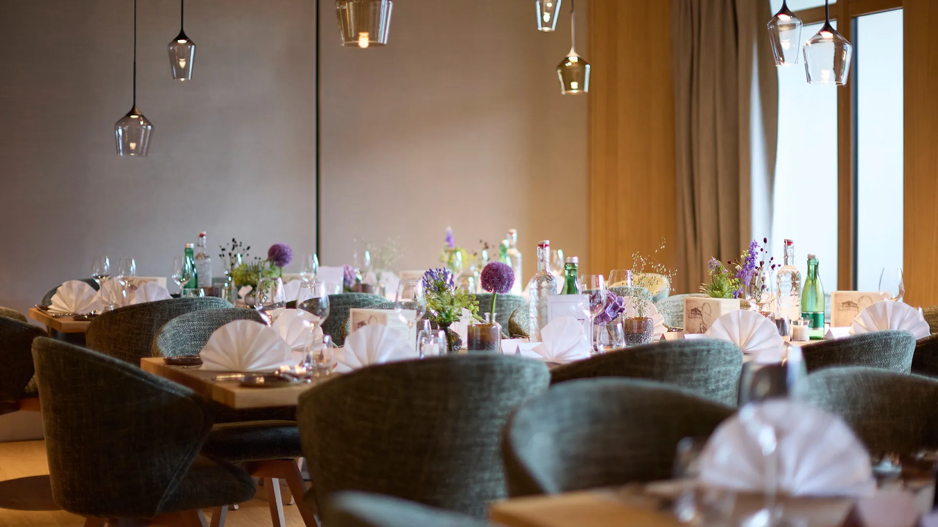 Restaurant mit festlich gedeckten Holztischen, grünen Stühlen, Blumenarrangements und modernen Pendelleuchten.