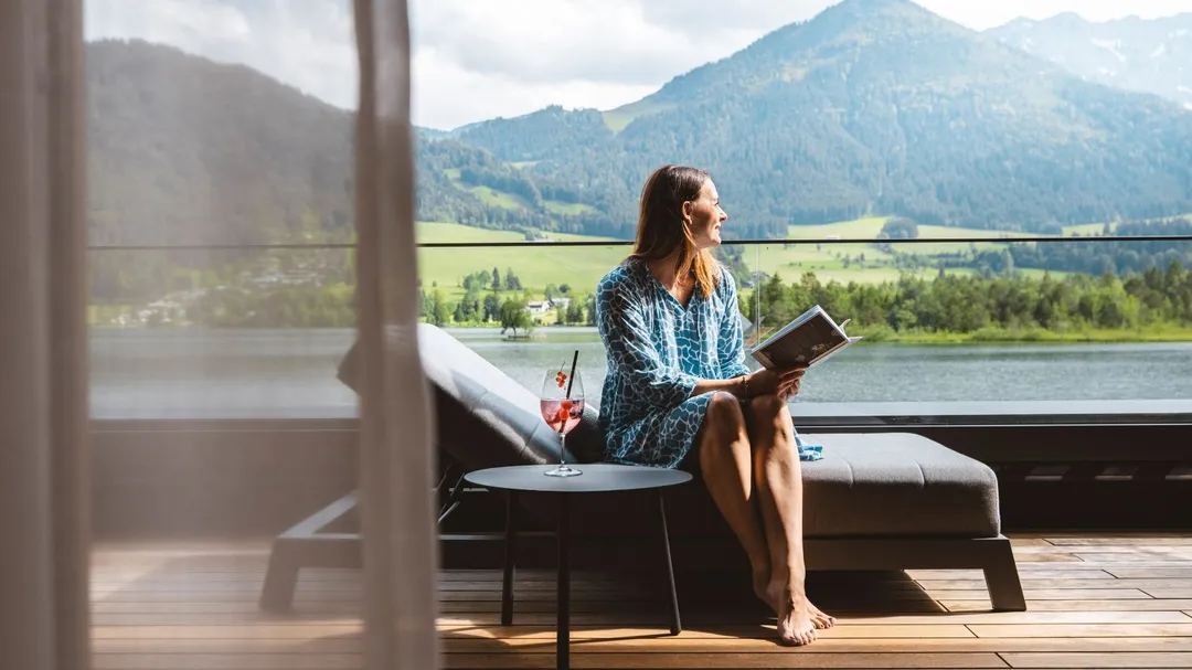 Frau auf Liege mit Getränk und Buch, Seeblick und Berge im Hintergrund