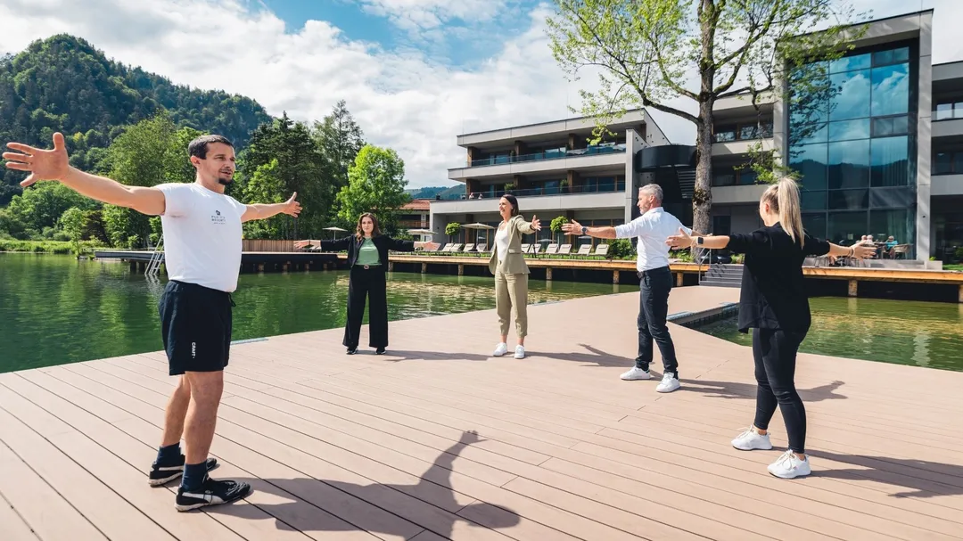 Personen mit ausgestreckten Armen auf Holzsteg am See vor Hotelgebäude