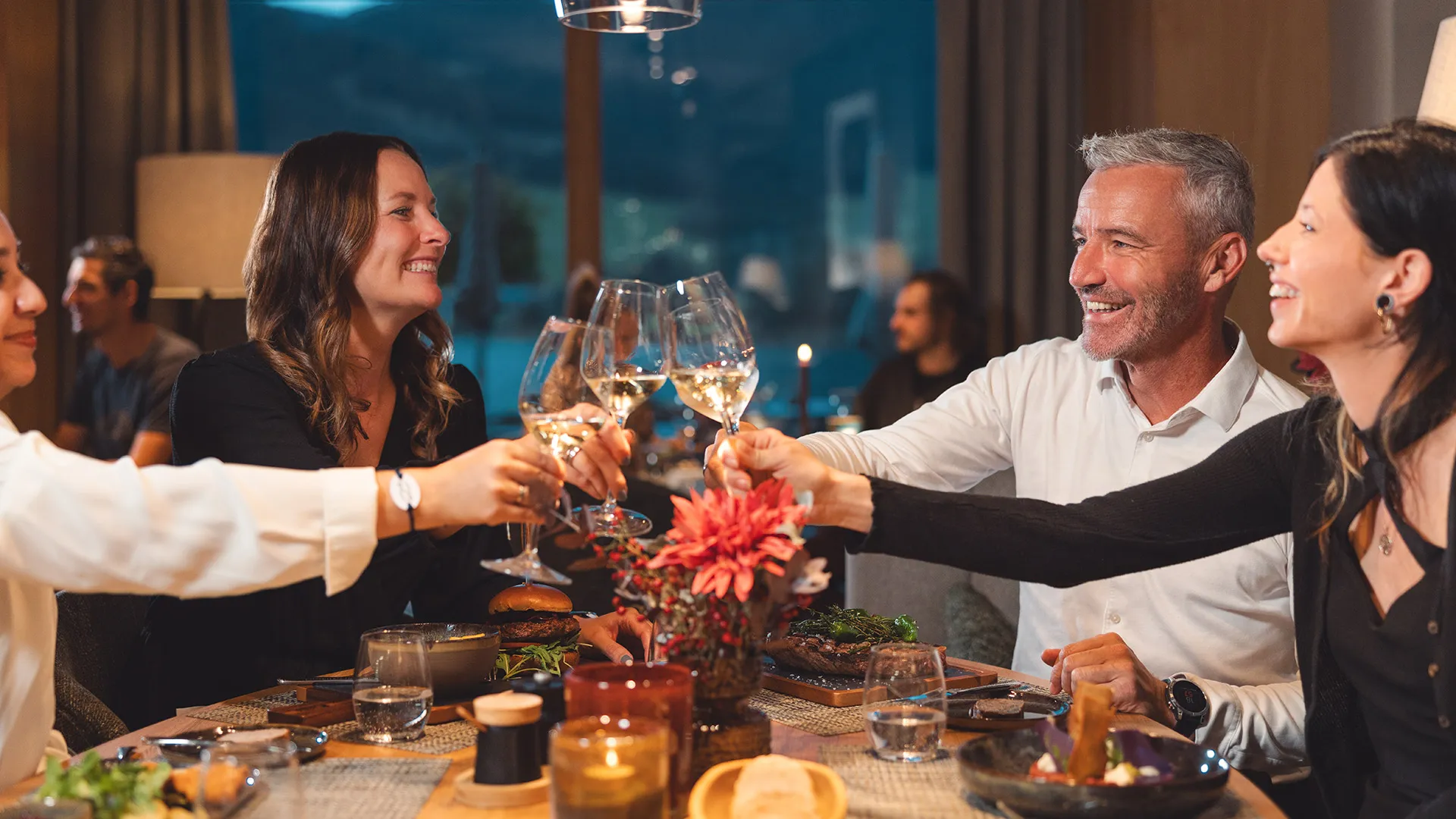 Eine Gruppe fröhlicher Menschen stößt beim Abendessen mit Weingläsern an einem festlich gedeckten Tisch an.