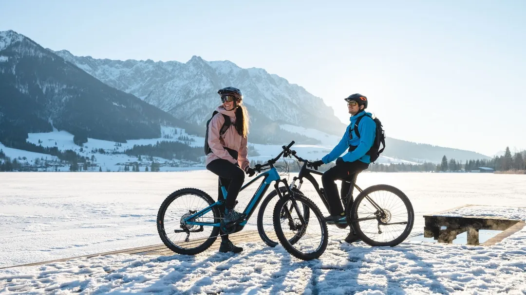 Paar mit Mountainbikes am zugefrorenen See vor verschneiter Bergkulisse