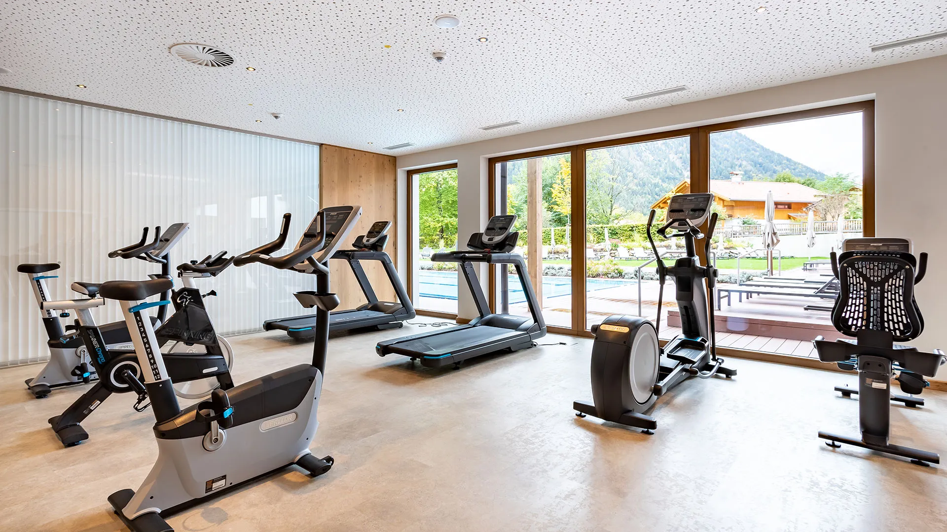 Fitnessraum mit Laufbändern und Fahrrädern, Blick auf Pool und Berge