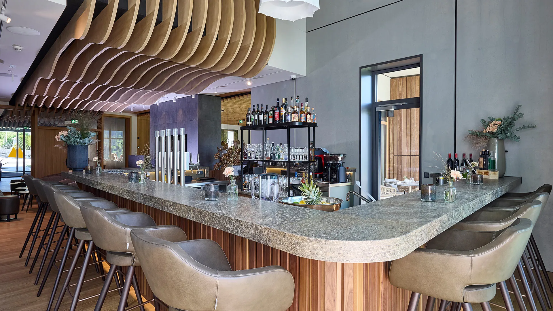 Moderne Hotelbar mit Steintheke, eleganten Barhockern, welliger Holzdecke und Flaschenregal.