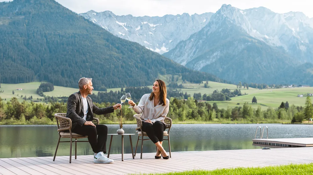 Ein Paar genießt Wein auf einer Holzterrasse am See, mit grünen Wäldern und schneebedeckten Bergen im Hintergrund.