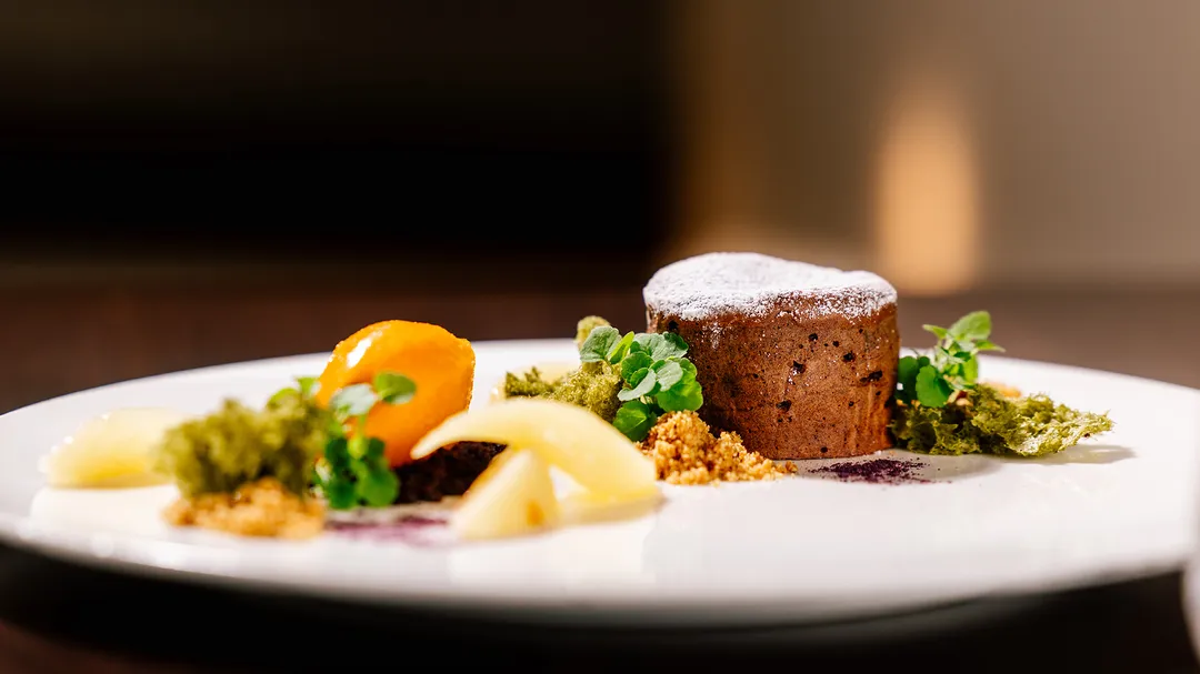 Schokoladendessert mit Puderzucker, Fruchtstück und Kräuterdeko auf weißem Teller