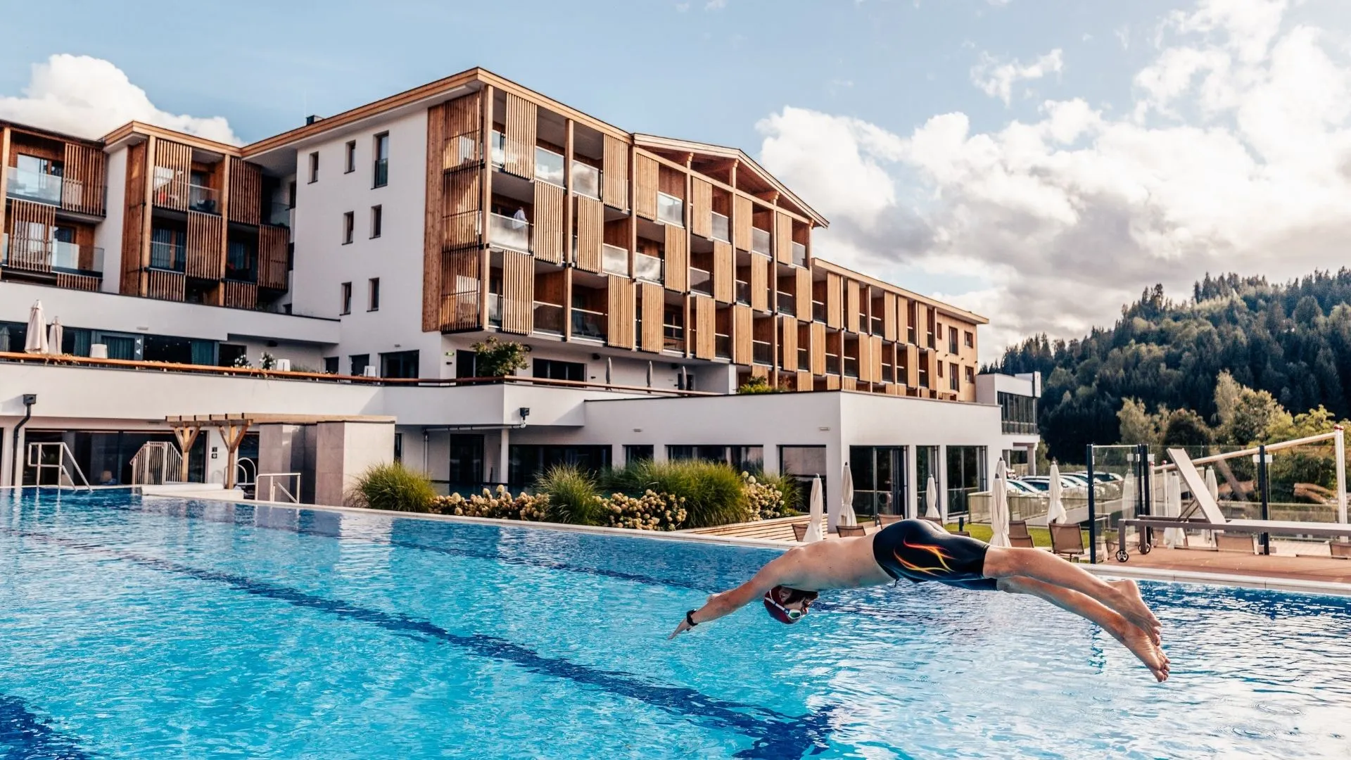 Mann springt in Außenpool vor Hotelgebäude mit Holzfassade