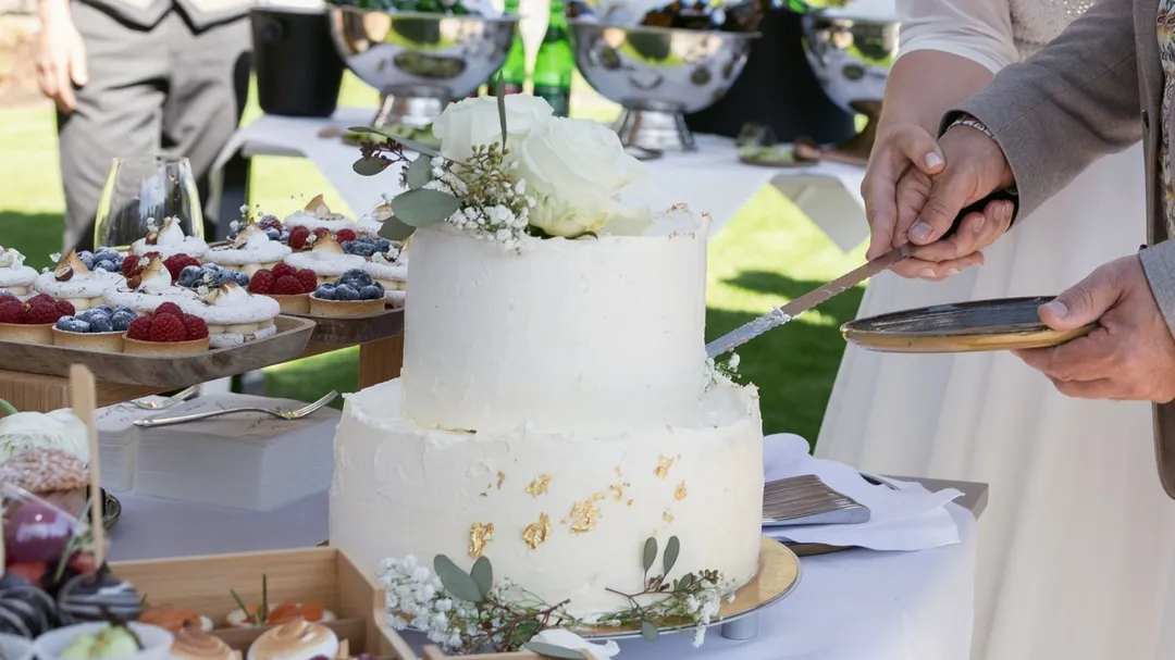 Zweistöckige Hochzeitstorte mit Blumen wird angeschnitten, im Hintergrund Dessertbuffet