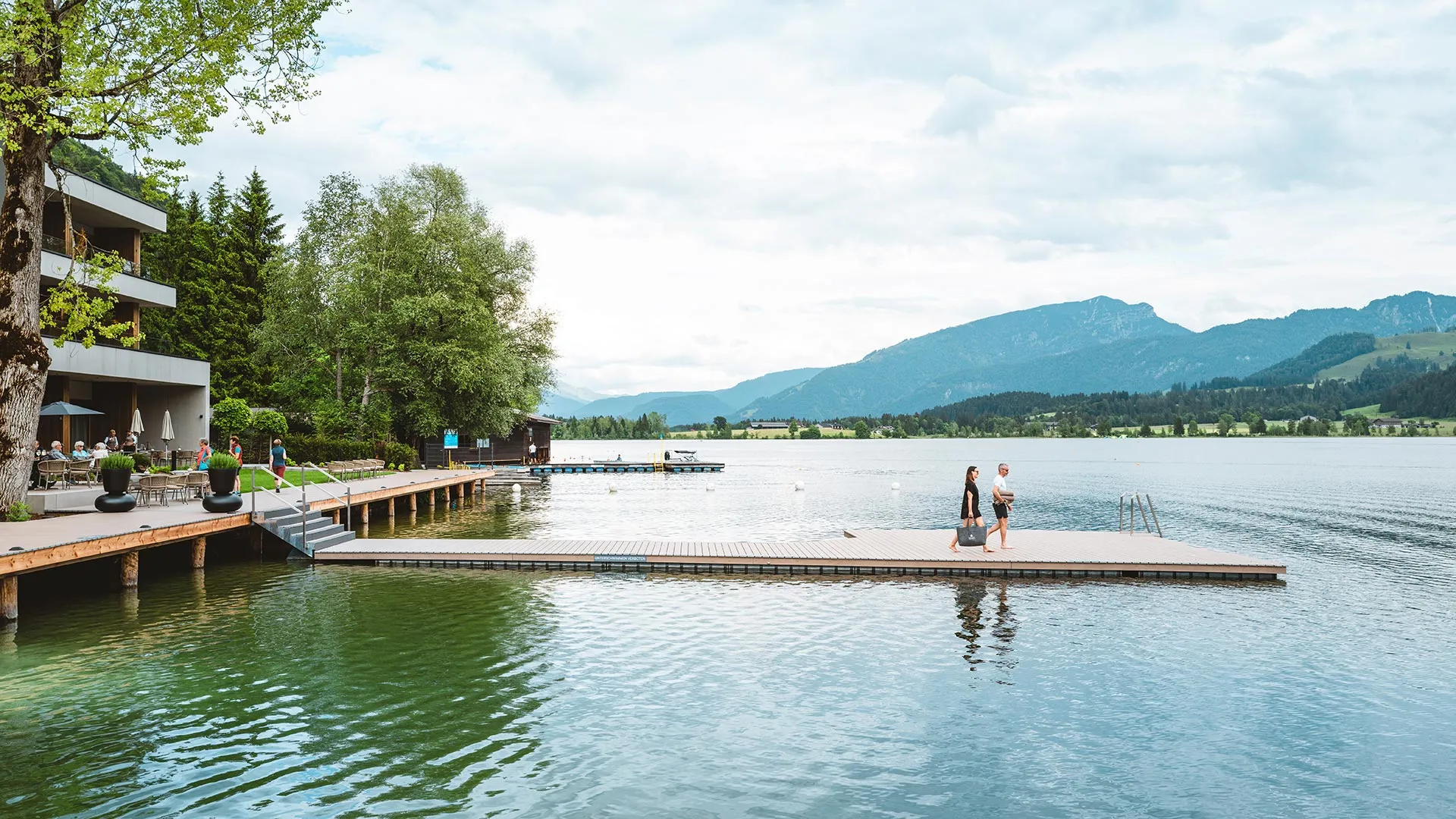 Modernes Hotel am Seeufer mit Holzsteg und Restaurantterrasse, umgeben von Bäumen und Bergen. Zwei Personen gehen auf einem Schwimmsteg.
