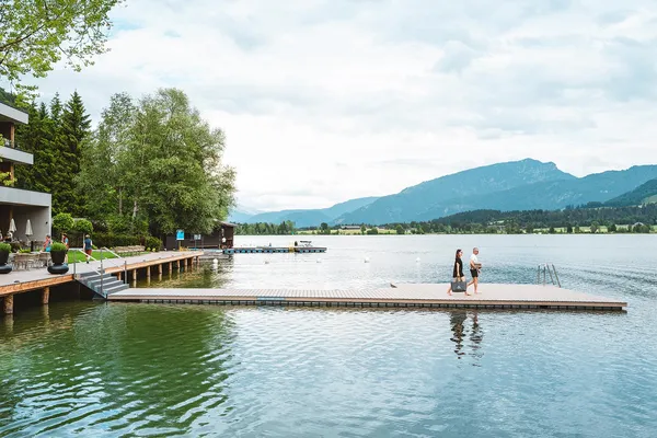Modernes Hotel am Seeufer mit Holzsteg und Restaurantterrasse, umgeben von Bäumen und Bergen. Zwei Personen gehen auf einem Schwimmsteg.
