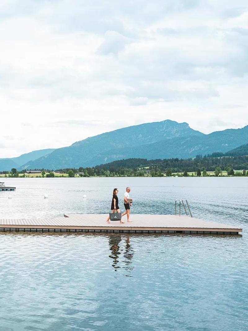 Modernes Hotel am Seeufer mit Holzsteg und Restaurantterrasse, umgeben von Bäumen und Bergen. Zwei Personen gehen auf einem Schwimmsteg.