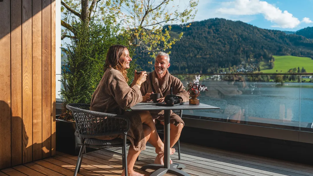 Ein Paar in Bademänteln sitzt lächelnd auf einem Balkon und trinkt Kaffee mit Blick auf einen See und Berge.