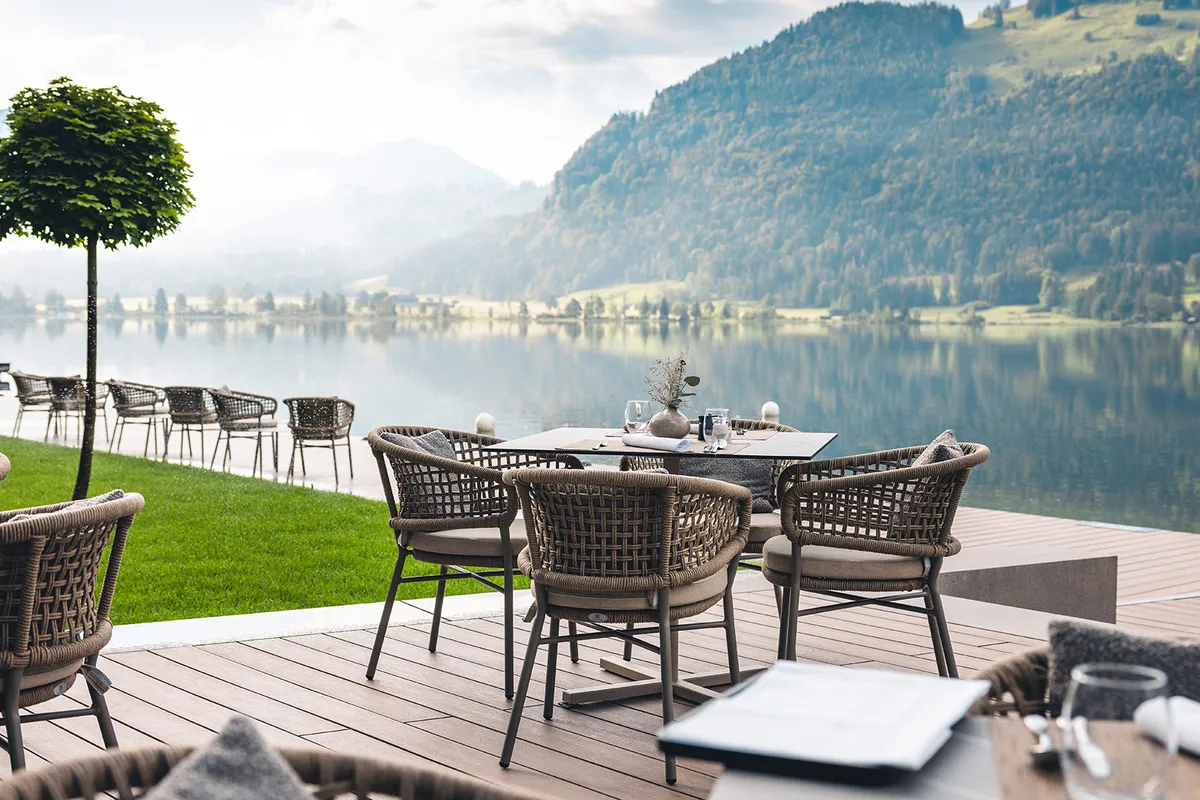 Stilvolle Restaurantterrasse am Seeufer mit gedeckten Tischen, Flechtstühlen und malerischer Bergkulisse im Hintergrund.