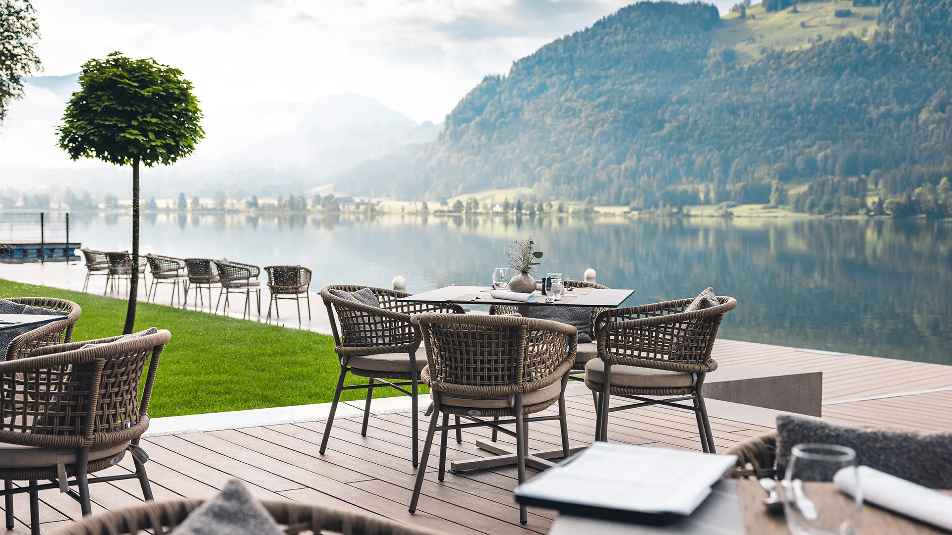 Stilvolle Restaurantterrasse am Seeufer mit gedeckten Tischen, Flechtstühlen und malerischer Bergkulisse im Hintergrund.