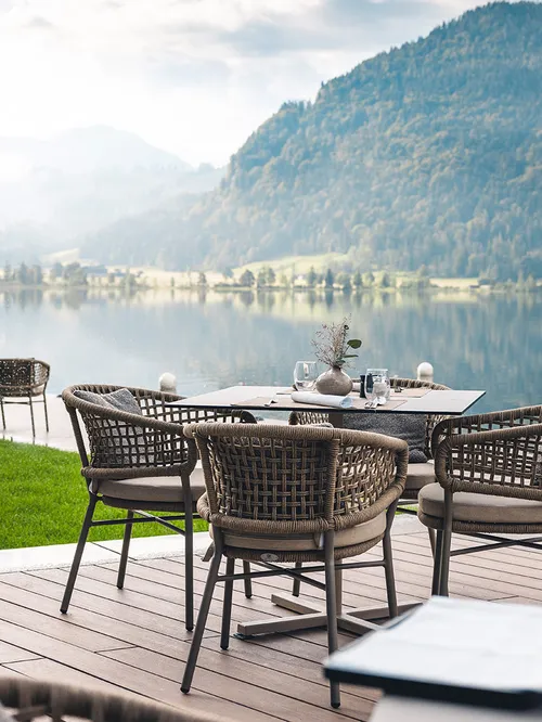 Stilvolle Restaurantterrasse am Seeufer mit gedeckten Tischen, Flechtstühlen und malerischer Bergkulisse im Hintergrund.