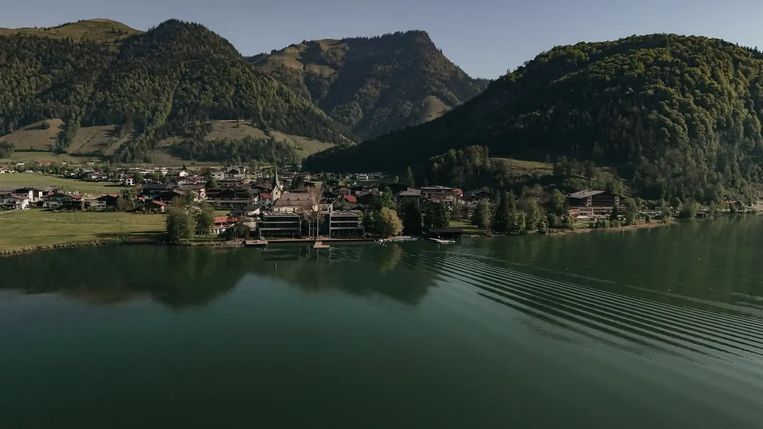 Dorf am See, umgeben von grünen Bergen, mit Wellen auf dem Wasser unter klarem Himmel.