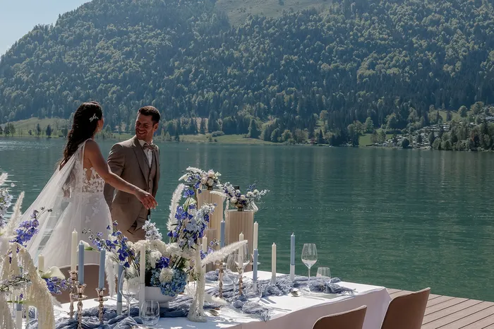 Glückliches Brautpaar bei einer Hochzeitsfeier am See mit Bergen und einem festlich dekorierten Tisch.