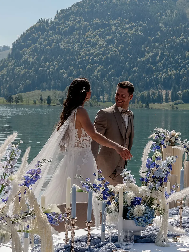 Glückliches Brautpaar bei einer Hochzeitsfeier am See mit Bergen und einem festlich dekorierten Tisch.