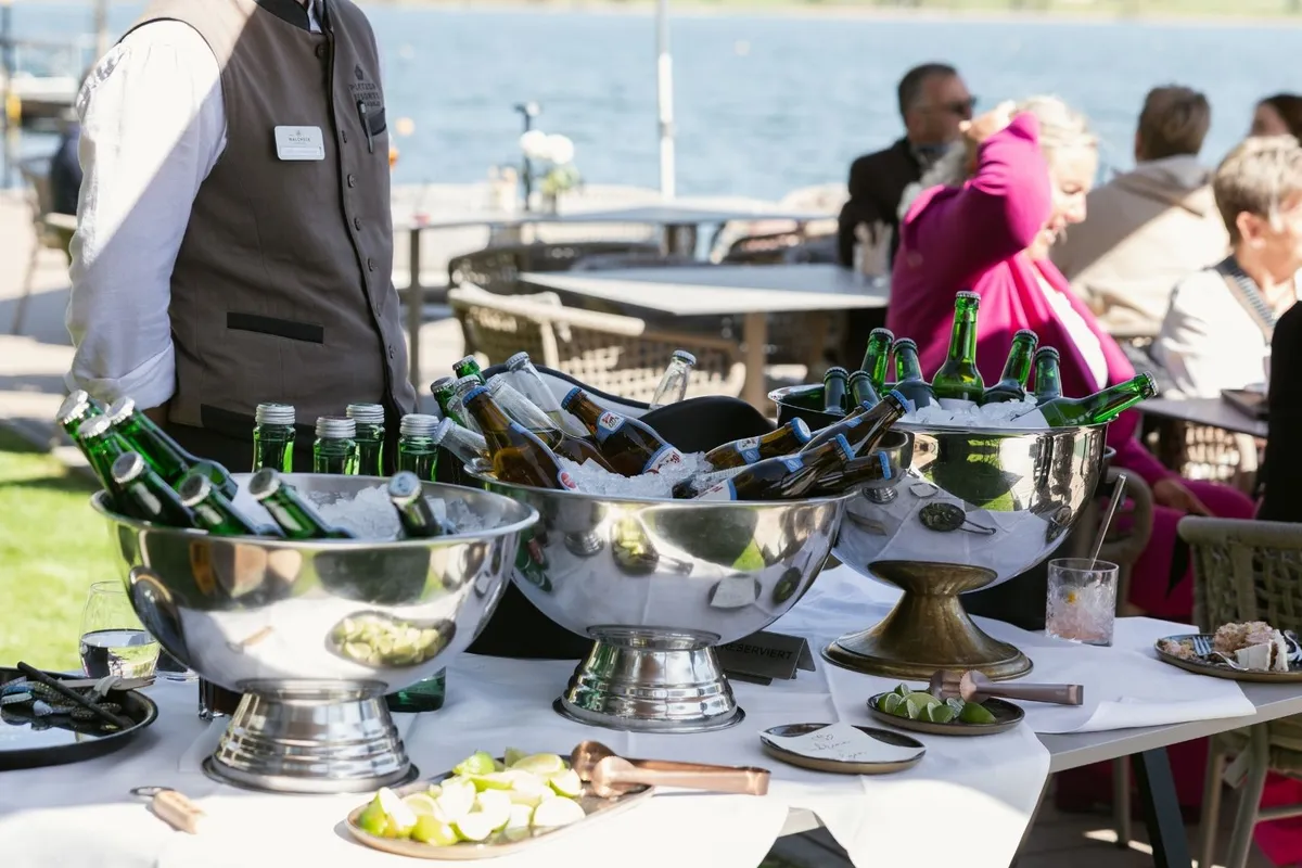 Bierflaschen in Eisschalen auf gedecktem Tisch am See mit Gästen im Hintergrund