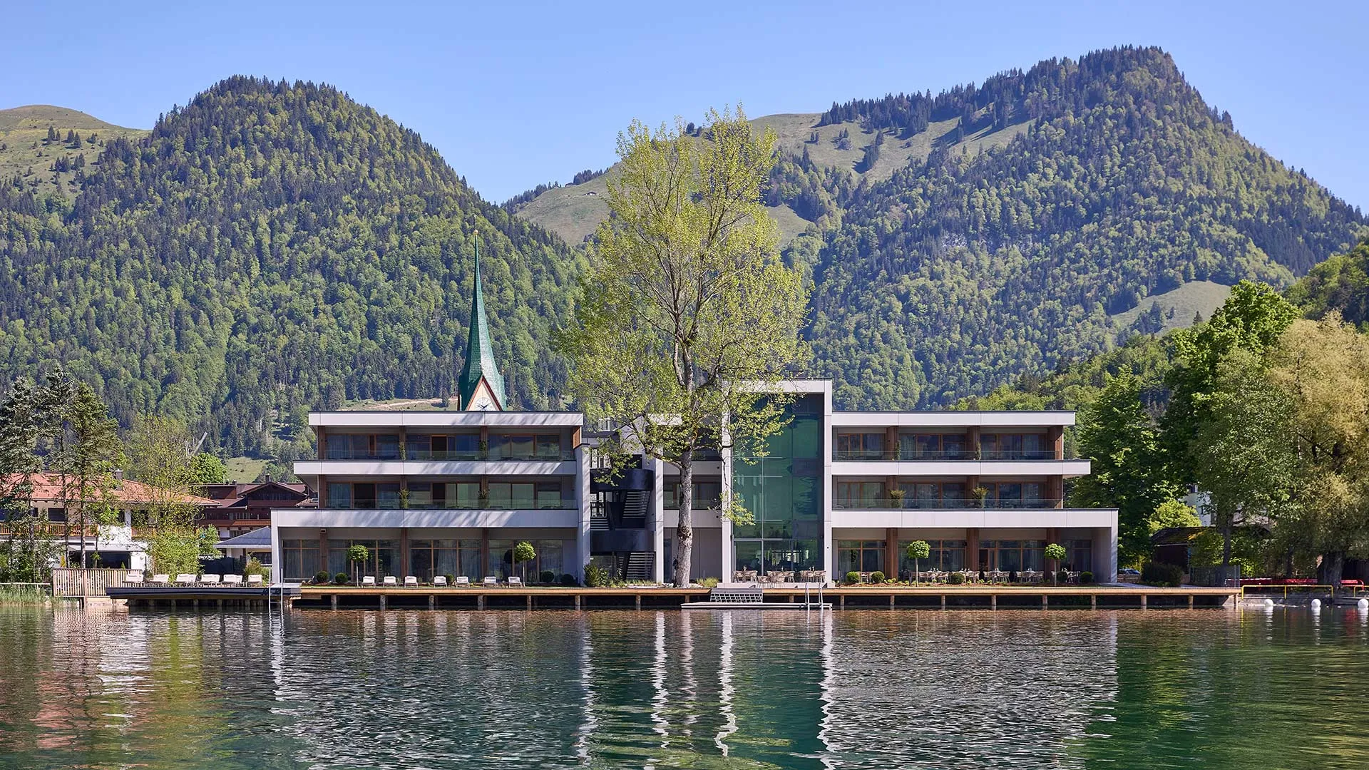 Modernes Gebäude am Seeufer mit einem Baum davor, einem Kirchturm links und bewaldeten Bergen im Hintergrund.