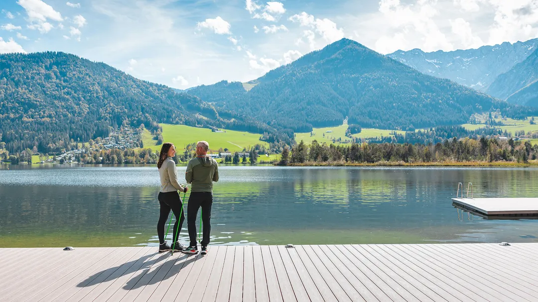 Paar mit Nordic-Walking-Stöcken an einem Steg am Walchsee vor Bergkulisse
