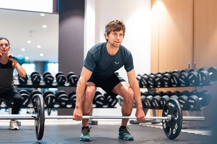 Mann hebt Langhantel im Fitnessraum, im Hintergrund Frau mit Kettlebells