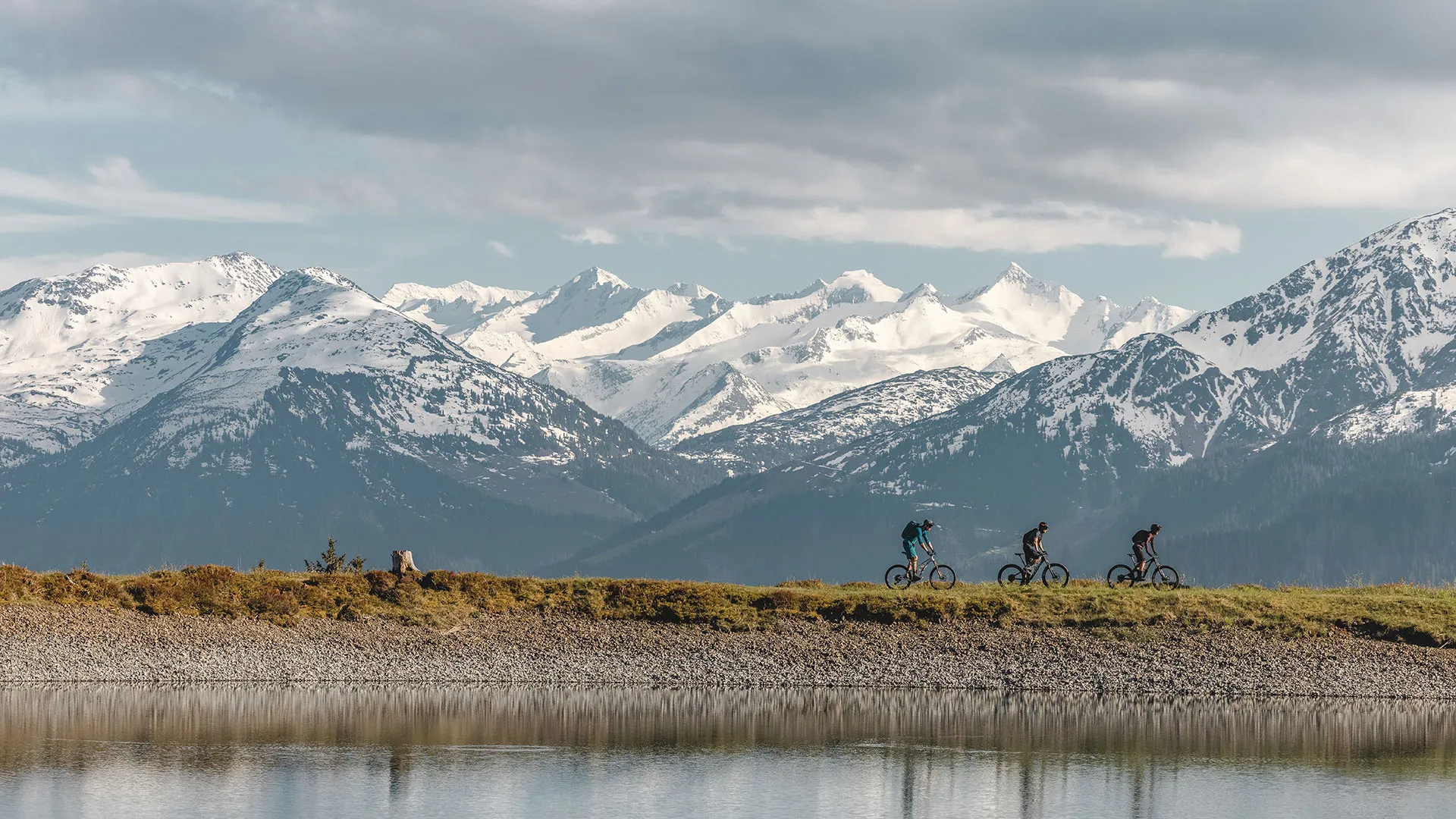 Mountainbiker fahren an einem See entlang, im Hintergrund schneebedeckte Berge