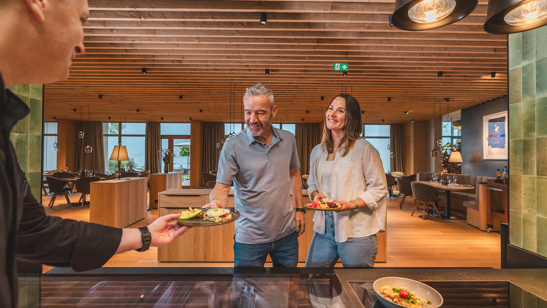Ein Kellner reicht einem lächelnden Paar Teller mit Frühstück in einem modernen Restaurant mit Holzelementen und Seeblick.