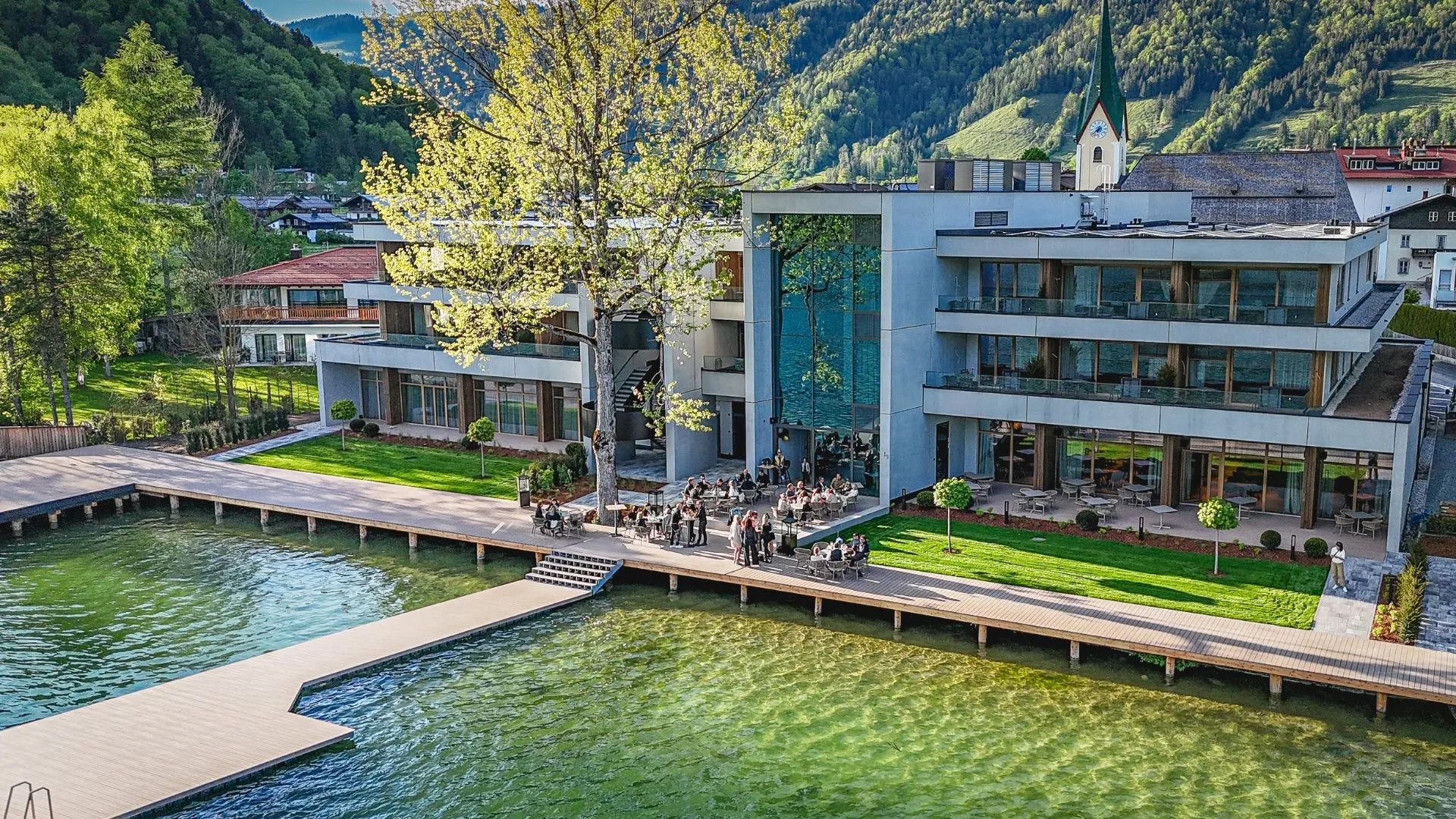 Hotel am See mit Terrasse und Steg vor bewaldeten Bergen