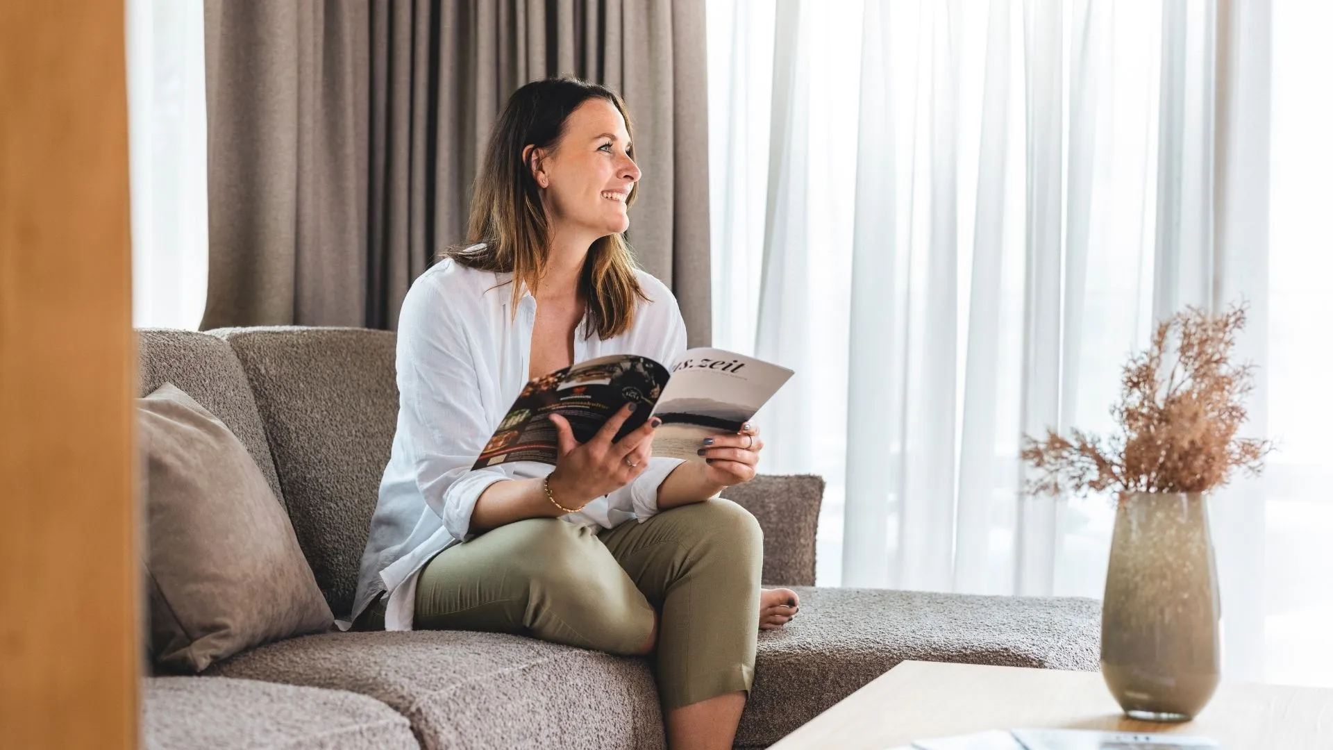 Frau sitzt lächelnd mit Magazin auf Sofa im Hotelzimmer