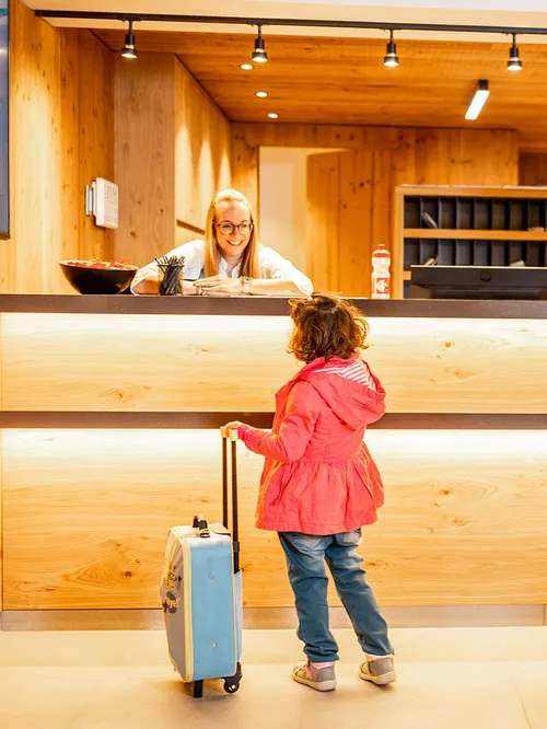 Kind mit Koffer an der Hotelrezeption, Mitarbeiterin lächelt hinter dem Empfangstresen