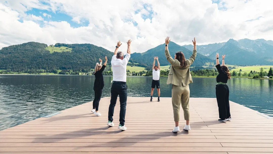 Gruppe macht Dehnübungen auf einem Steg am See vor Bergpanorama