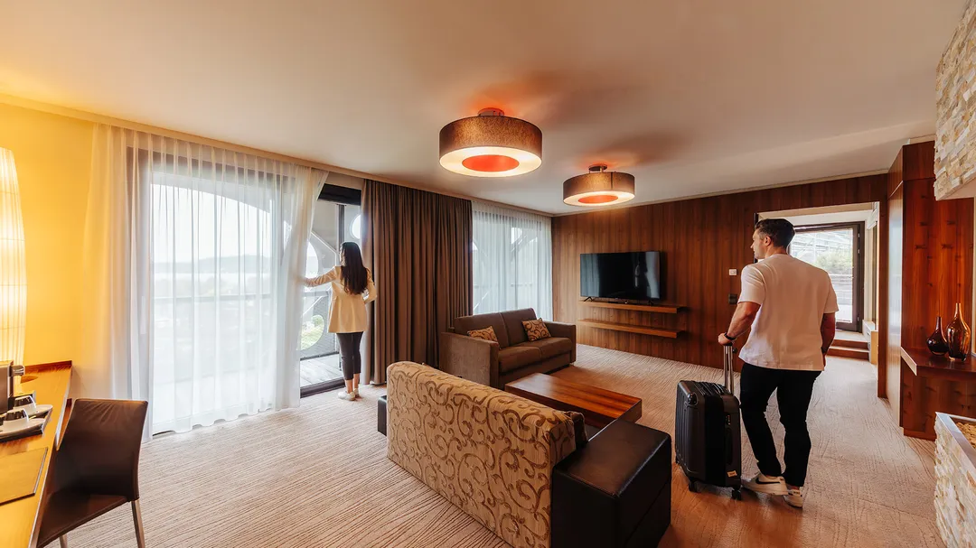 Paar bezieht Hotelsuite mit Koffer, Sofas und Vorhang zum Balkon
