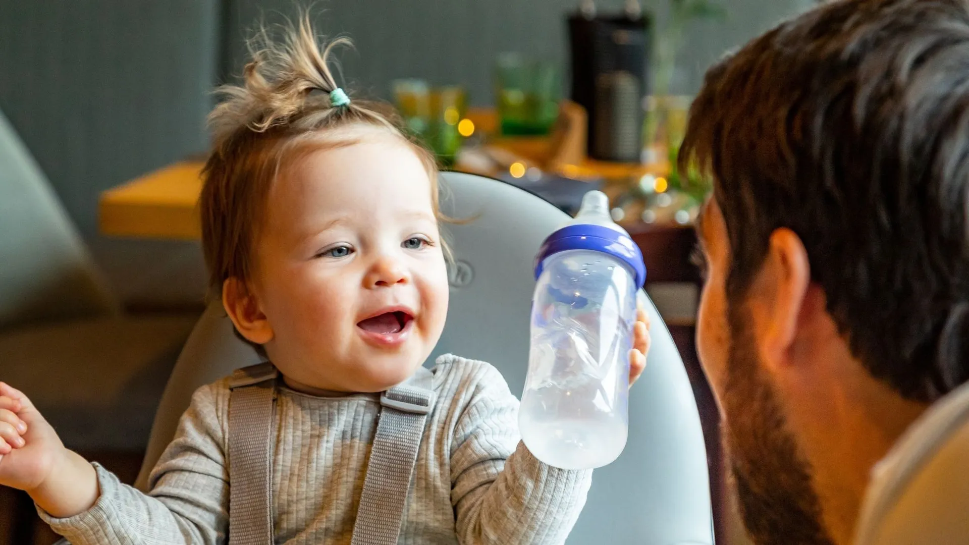Lachendes Baby im Hochstuhl hält eine Trinkflasche, Vater sitzt daneben
